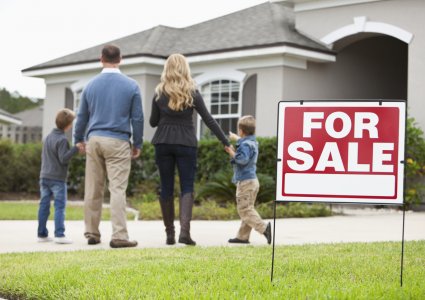 Family with two boys (4 and 6 years) standing in front of house with FOR SALE sign in front yard. Focus on sign.